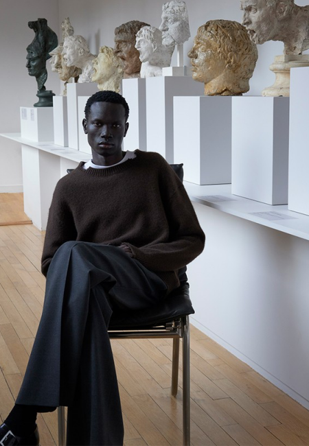 Sanat eserlerinin sergilendiği bir ortamda, Sandro gibi en ünlü erkek giyim markalarının Parizyen minimal stilini yansıtan koyu tenli bir model, kahverengi bol bir kazak, modern kesim koyu gri geniş paçalı pantolon ve toka detaylı siyah ayakkabılarla zamansız bir zarafet sunuyor.