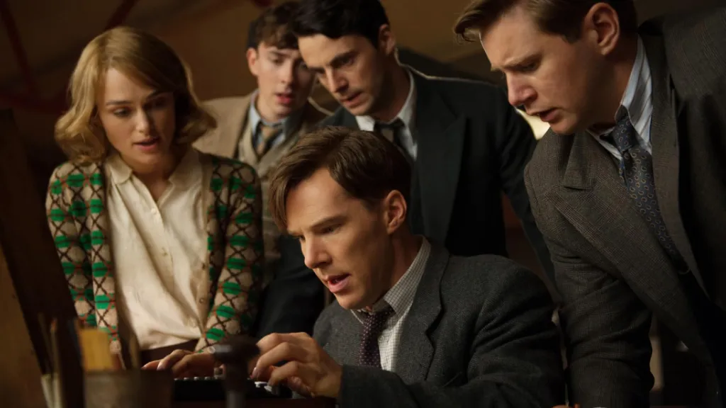 The Imitation Game (Enigma) filminden bir sahne: Alan Turing karakteri (Benedict Cumberbatch) eski bir makine üzerinde yoğun şekilde çalışırken, etrafındaki ekip arkadaşları heyecan ve merakla onu izliyor. Ortam, II. Dünya Savaşı dönemine ait bir şifre çözme laboratuvarını yansıtıyor.