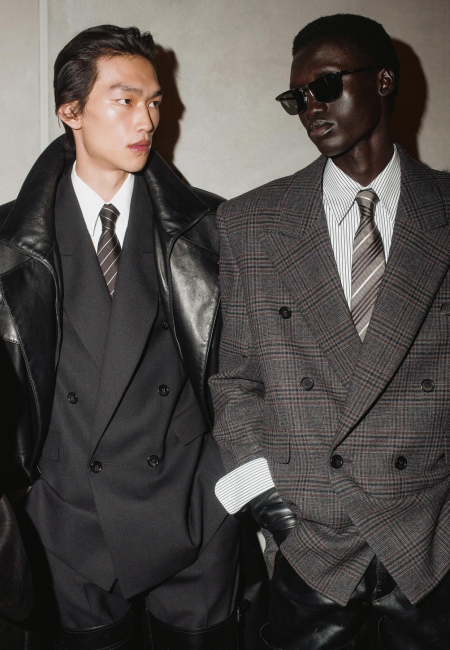 Siyah kruvaze takım elbise ve şık bir deri ceketle poz veren Asyalı bir model ile ekose blazer, çizgili gömlek ve güneş gözlüğüyle dikkat çeken koyu tenli başka bir modelin yan yana durduğu bu moda çekimi, en ünlü erkek giyim markalarının sunduğu lüks ve modern stil çeşitliliğini çarpıcı bir şekilde sergiliyor.