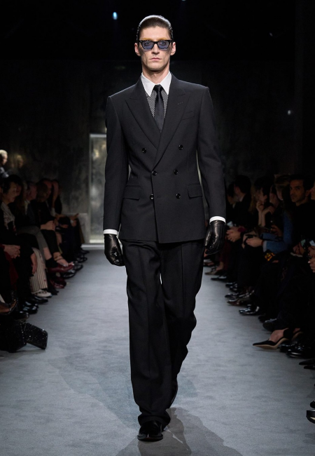 Bir podyumda, siyah kruvaze takım elbisesi, beyaz gömleği, desenli kravatı, siyah deri eldivenleri ve keskin koyu renk güneş gözlükleriyle yürüyen erkek model, Tom Ford gibi lüks bir erkek giyim markasının maskülen, sofistike ve kusursuz terzilik anlayışını modern bir şıklıkla sergiliyor.