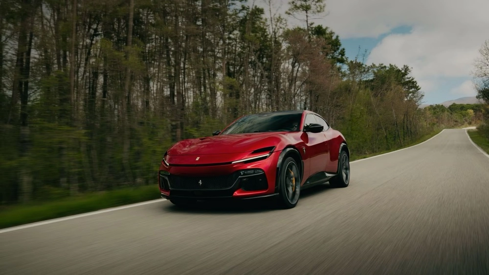 Kırmızı bir Ferrari Purosangue, etrafı ormanlık ağaçlarla çevrili kıvrımlı asfalt bir yolda dinamik bir şekilde ilerlerken, aracın çarpıcı ön tasarımını ve şık dış mekan detaylarını gözler önüne seriyor.