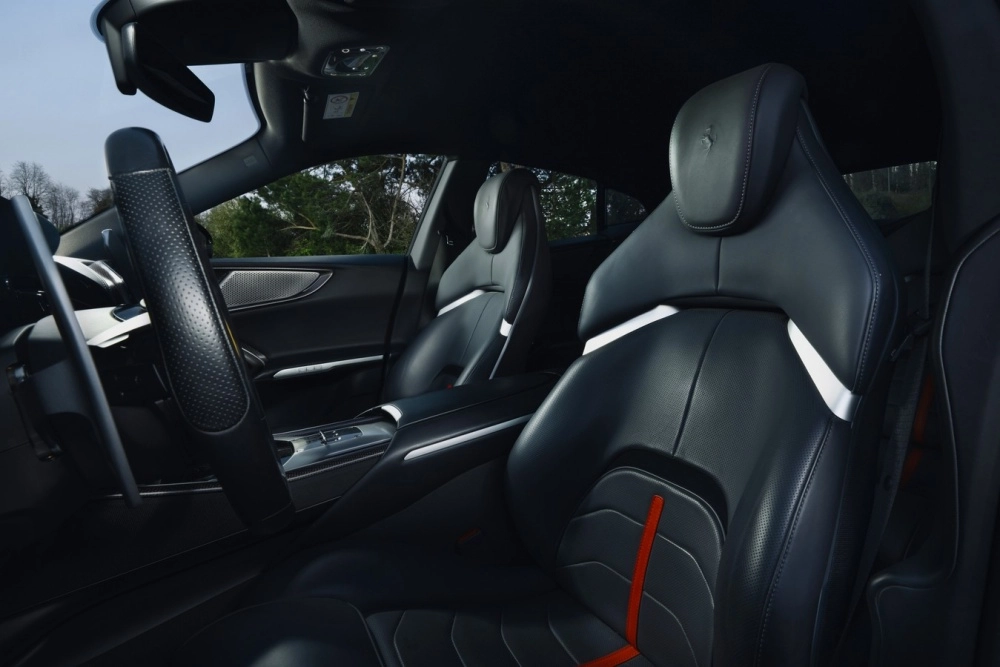 Ferrari Purosangue'nin şık siyah iç mekan tasarımını vurgulayan bu fotoğrafta, kırmızı çizgili ve beyaz yan destekli delikli deri ön koltuklar, modern direksiyon simidi ve minimalist orta konsol ön planda.
