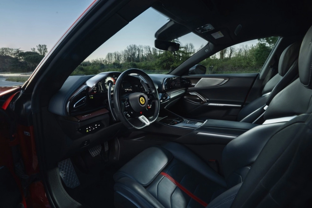 Ferrari Purosangue'nin modern siyah iç mekan tasarımını geniş açıyla gösteren bu görselde, sürücü tarafındaki direksiyon simidi, dijital gösterge paneli ve şık ön konsol detayları belirginleşirken, geniş ön camdan dışarıdaki yeşilliklerle çevrili yol manzarası seçiliyor.