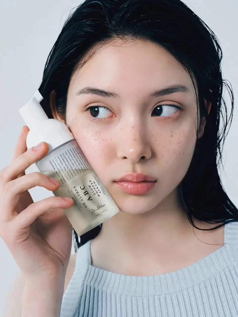 Ortadan ayrılmış ıslak saçları ve çilleri olan genç bir kadın, açık mavi nervürlü bir atlet giyerek Dr. Althea ABC Glow Whipped Serum şişesini hafifçe sol yanağına yaslamış, yan tarafa bakarken Fiddy Metodu ile ilişkili bir cilt bakım ürününü tanıtıyor.