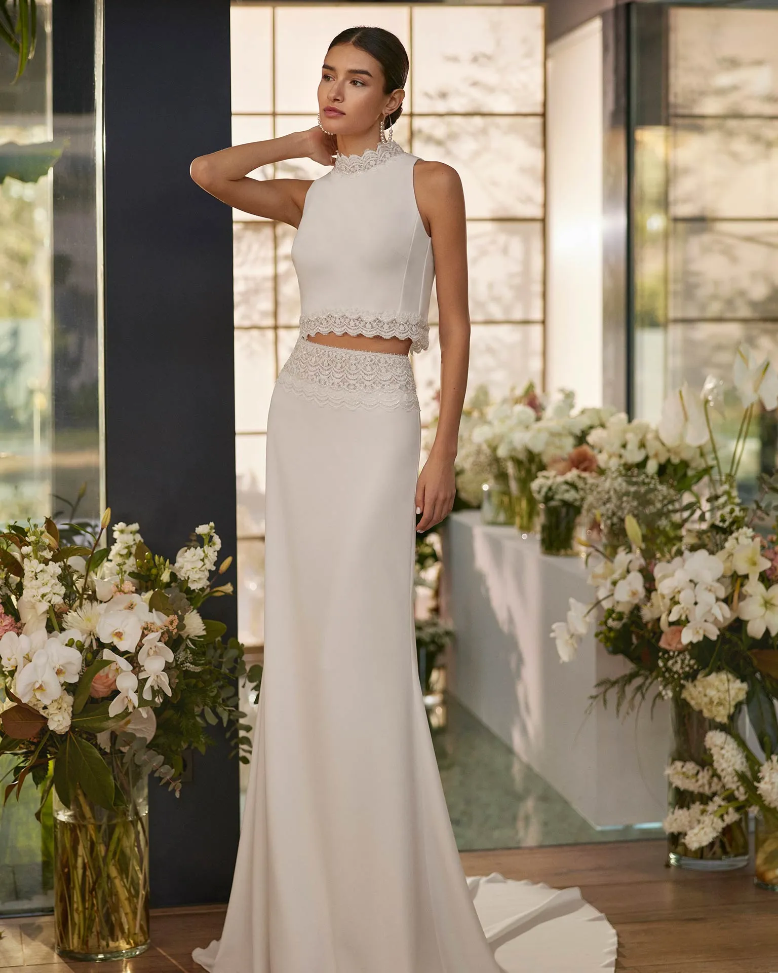 Rosa Clará iki parçalı modern gelinlik; yüksek yaka dantel işlemeli kolsuz crop top ve dantel bel detaylı, sade ve akışkan maksi etek.