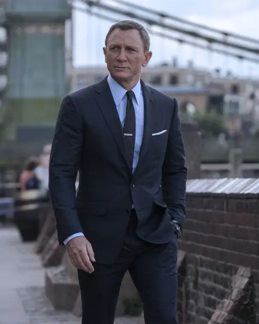 Daniel Craig, James Bond karakteriyle kusursuz kesimli koyu renk takım elbisesi, açık mavi gömleği, kravat iğneli koyu renk kravatı ve beyaz mendiliyle şehrin modern dokusunda yürürken, postta ele alınan James Bond stilinin sofistike ve zamansız örneklerinden birini sergiliyor.