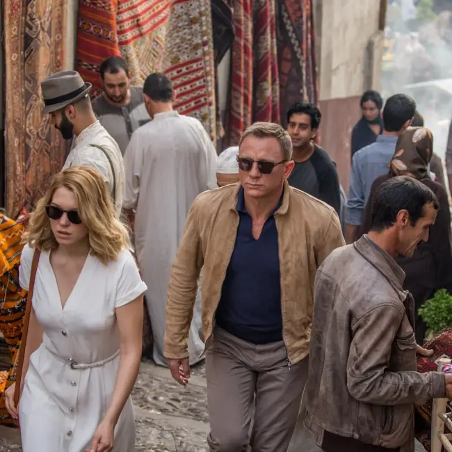 James Bond, kum rengi süet ceketi, lacivert polo tişörtü ve güneş gözlükleriyle, beyaz elbiseli bir kadınla birlikte, arkasında geleneksel dükkanların sıralandığı kalabalık bir sokakta, günlük yaşamda da sürdürdüğü ikonik James Bond stilini ve zahmetsiz şıklığını yansıtıyor.
