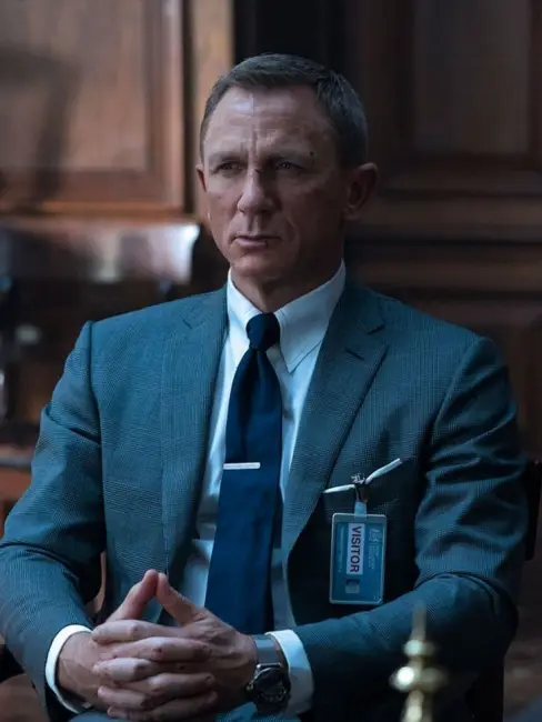 Gri renkli, kusursuz kesimli bir takım elbise, beyaz gömlek, lacivert kravat ve zarif bir kravat iğnesi ile Daniel Craig, James Bond stili içinde kollarını kavuşturmuş, sol bileğinde şık bir saat ve yakasında ziyaretçi kartı ile ciddi bir ifadeyle oturuyor.