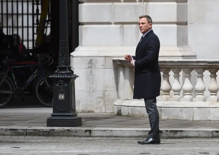 Daniel Craig, James Bond stilinin zamansız zarafetini kusursuzca yansıtan, diz hizasında koyu renk şık bir palto, gri takım elbise pantolonu ve klasik deri botlarıyla şehir ortamında sofistike bir duruş sergiliyor.