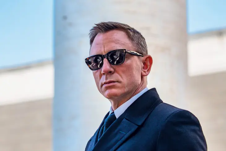 James Bond stilinin ikonik bir öğesi olan, koyu asetat çerçeveli güneş gözlükleri takmış, lacivert bir takım elbise veya palto içinde, kararlı ve gizemli bir ifadeyle doğrudan bakan Daniel Craig, sofistike ve zamansız imajıyla dikkat çekiyor.