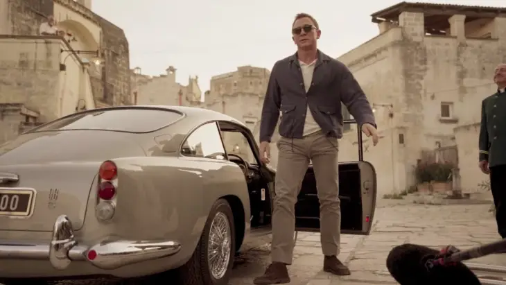 James Bond stilinin vazgeçilmez bir parçası olan klasik Aston Martin otomobilinden, taş binalarla çevrili bir sokakta inerken güneş gözlükleri ve spor ceketiyle görüntülenen Daniel Craig, karakterin zamansız ve karizmatik duruşunu sergiliyor.