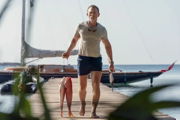 Yalınayak Daniel Craig, açık renk tişört ve koyu renk şortuyla ahşap iskelede karizmatik bir duruş sergileyerek yürürken, bir elinde tuttuğu iki kırmızı balık ve diğer elindeki zıpkınla James Bond stilinin gündelik şıklığını ve fonksiyonel tarafını yansıtıyor.