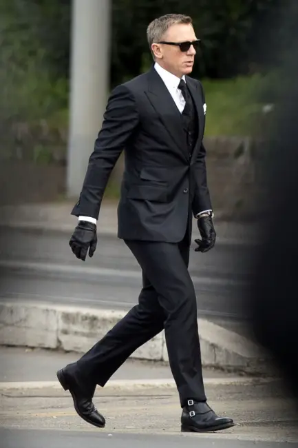 Daniel Craig, James Bond rolünde, koyu renk üç parçalı bir takım elbise, beyaz gömlek, kravat, güneş gözlüğü, siyah deri eldivenler ve çift tokalı deri botlarla karizmatik ve kusursuz James Bond stiline uygun sofistike bir şekilde yolda yürüyor.