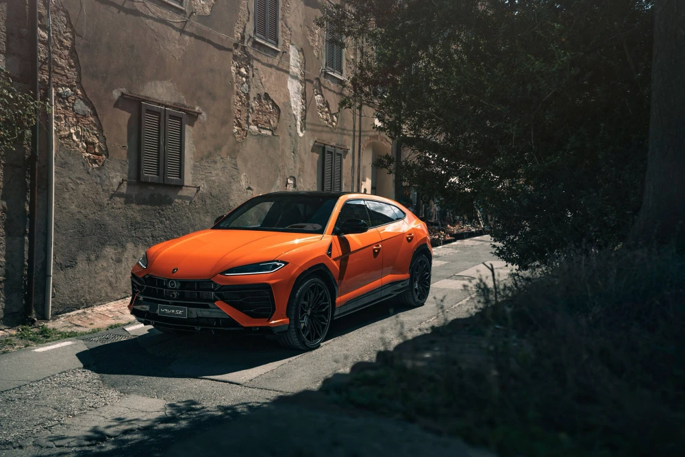 Turuncu renkteki, şık dış mekan tasarımına sahip Lamborghini Urus SE Plug-in Hybrid lüks SUV, güneşli bir Toskana kasabasının dar taş döşeli sokağında, eski ve yıpranmış taş binalarla yeşil ağaçların gölgeleri arasında park halinde görülüyor.