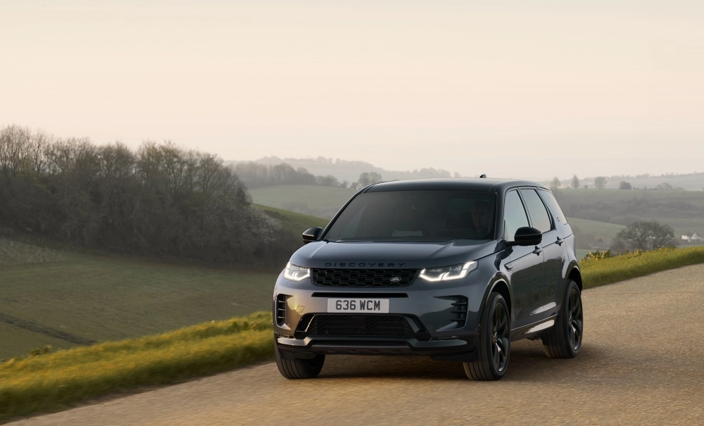 Land Rover Discovery Sport PHEV modelinin dış tasarımı, farları açık koyu renkli şarj edilebilir hibrit SUV aracın yeşil tepeler ve ağaçlarla çevrili hafif eğimli bir yolda dinamik bir açıyla konumlandığını gösteriyor.