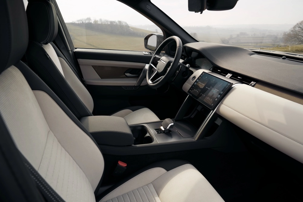 Land Rover Discovery Sport PHEV şarj edilebilir plug-in hibrit SUV modelinin ön kabinindeki koyu ve açık renk kombinasyonlu koltuklar, modern dokunmatik bilgi-eğlence ekranı ve dışarıdaki geniş manzarayı yansıtan şık iç mekan tasarımı.