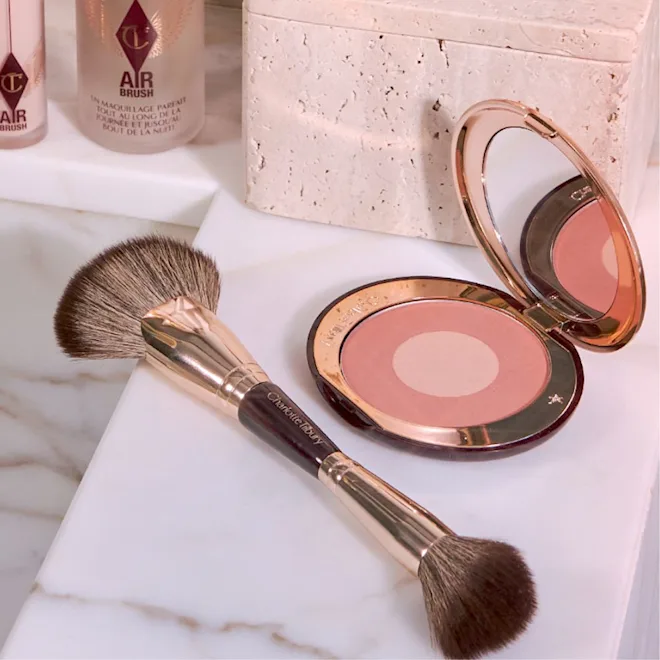 Mermer bir yüzeyde, altın renkli detaylara sahip, farklı uygulamalar için iki ucu bulunan Charlotte Tilbury makyaj fırçası, ortası açık pembe, dışı koyu pembe tonlarında allık içeren ve aynası açık duran kompakt bir paletle birlikte gösterilirken, arka planda Airbrush serisinden fondöten şişeleri de seçilebiliyor.