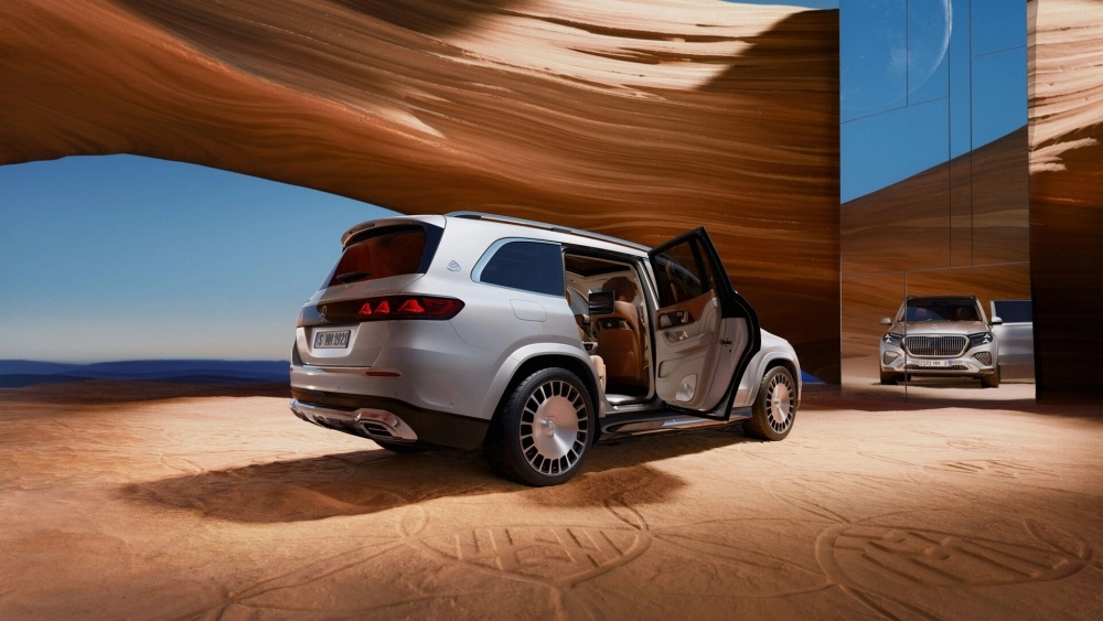 Açık renkli lüks bir Mercedes-Maybach GLS SUV, kumlu ve dalgalı kaya oluşumlarıyla çevrili bir arazide, arka yolcu kapıları açıkça görünen kahverengi deri döşemeli arka koltuk makam konforunu ve şık dış mekan tasarımını gözler önüne seriyor. Yansıtıcı yüzeyde aracın ön görünümü ve Ay detaylı gökyüzü belirginleşiyor.
