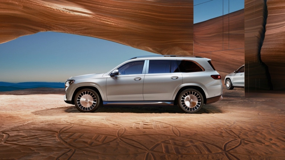 Gümüş renkli Mercedes-Maybach GLS lüks SUV, kumlu ve üzerinde desenler bulunan bir zeminde yan profilinden sergileniyor; aracın şık dış mekan tasarımı, arka plandaki dalgalı kahverengi kaya oluşumları ve yansıtıcı yüzeylerle birleşen açık mavi gökyüzüyle dikkat çekiyor.