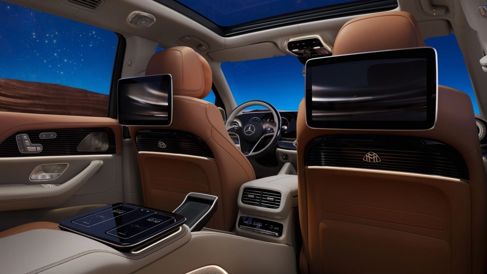 Mercedes-Maybach GLS'in lüks iç mekan tasarımını detaylandıran bu görselde, kahverengi deri arka koltuklardan ön tarafa doğru bakıldığında, başlık arkalarında konumlanan multimedya ekranları, orta konsoldaki gelişmiş kontrol paneli ve yan camdan görünen yıldızlı gece manzarası, makam konforunu en üst düzeye çıkarıyor.
