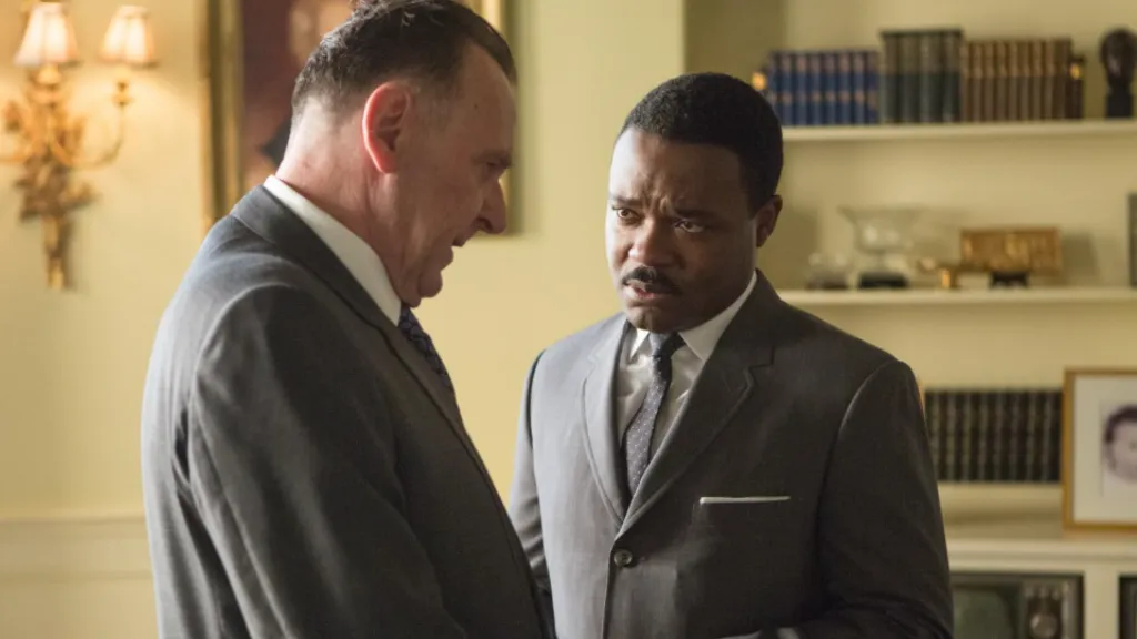 Selma (Özgürlük Yürüyüşü) filminden bir sahne: Martin Luther King Jr.'ı canlandıran karakter, ciddi bir ifadeyle bir adamla yüz yüze konuşuyor. İkisi de takım elbise giymiş, arka planda kitaplık ve eski tip bir televizyon yer alıyor. Sahne, siyasi ve toplumsal bir karar anını yansıtıyor.