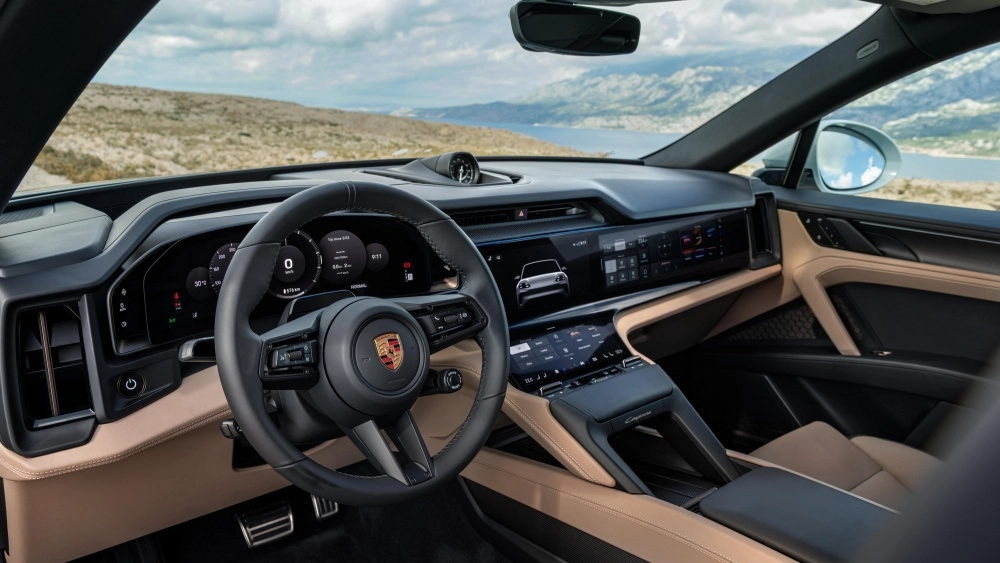 Porsche Cayenne elektrikli SUV'nin iç mekan tasarımı, bej döşemeleriyle öne çıkan modern ön konsolunu, çoklu dijital ekranları ve şık direksiyon simidini gösterirken, ön camdan görünen dağlık arazi ve deniz manzarasıyla lüks ve ferah bir atmosfer sunuyor.