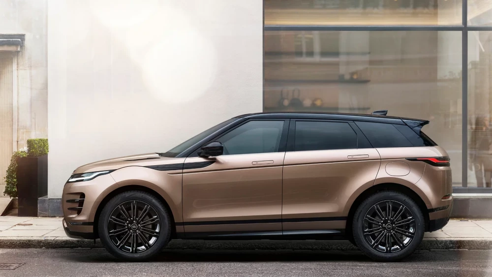 Modern şehir mimarisi önünde, şampanya metalik bej gövdesi ve kontrast oluşturan siyah tavanıyla dikkat çeken Range Rover Evoque şarj edilebilir hibrit kompakt SUV otomobil, kaldırım kenarına park edilmiş bir yan profil görünümü sunuyor.