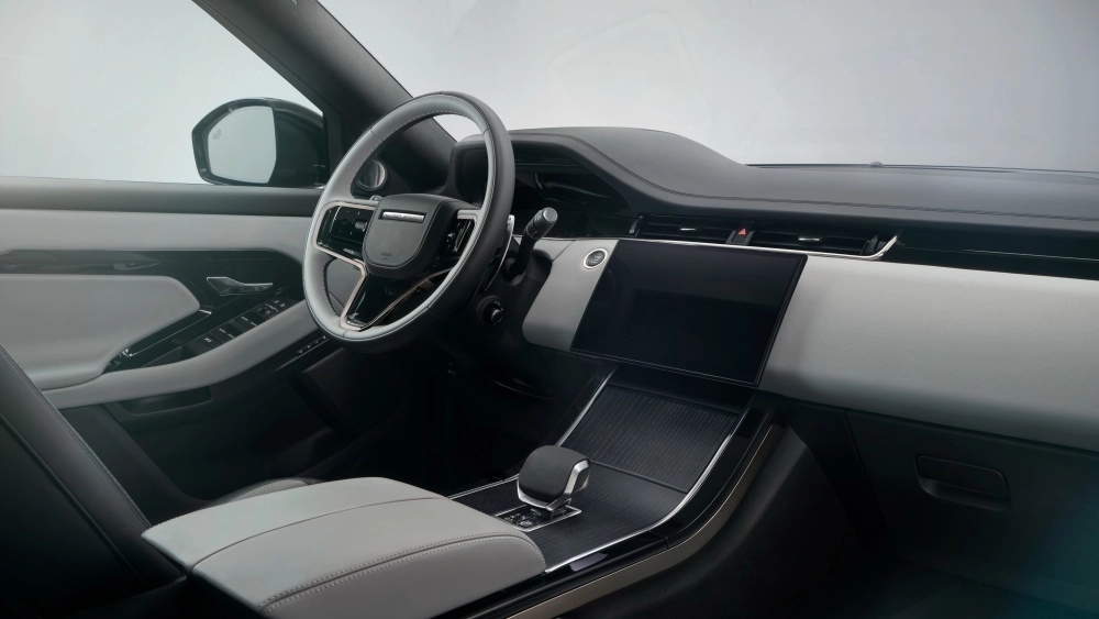 Range Rover Evoque Phev şarj edilebilir hibrit kompakt SUV otomobilin iç mekan tasarımı, açık gri ve siyah tonlarının hakim olduğu modern bir estetikle, şık direksiyon simidi, merkezi dokunmatik bilgi-eğlence ekranı ve vites kolunu detaylıca gösteriyor.