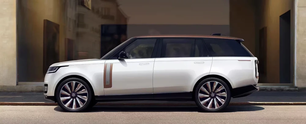 Yüksek performanslı bir lüks SUV olan beyaz Range Rover SV'nin kahverengi tavanı ve yanındaki zarif şerit detaylarıyla dikkat çeken modern tasarımı, çok kollu parlak jantlarıyla şık bir şekilde bir şehir caddesinde sergileniyor.
