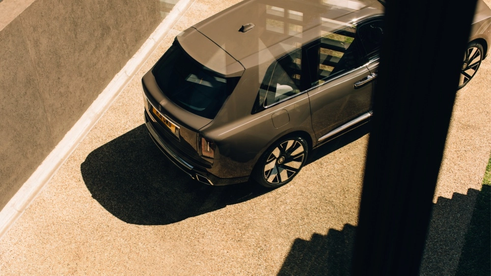Güneşli bir ortamda, açık kahverengi çakıllı bir zeminde park edilmiş, koyu bej renkte Rolls-Royce Cullinan Series 2 lüks SUV'un arka tasarımı, dinamik jantları ve yan profili üstten kuşbakışı açıyla detaylı bir şekilde görünüyor.