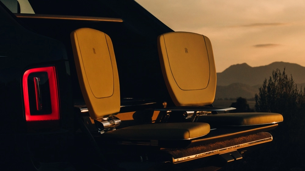 Rolls-Royce Cullinan lüks SUV'un açık bagajından dışarı doğru uzanan, markanın ikonik logosunu taşıyan bej deri piknik koltukları, sol arka lambasının kırmızı ışığı ve gün batımının vurduğu dağlık bir manzarayla birleşerek şık bir dış mekan konforu sunuyor.