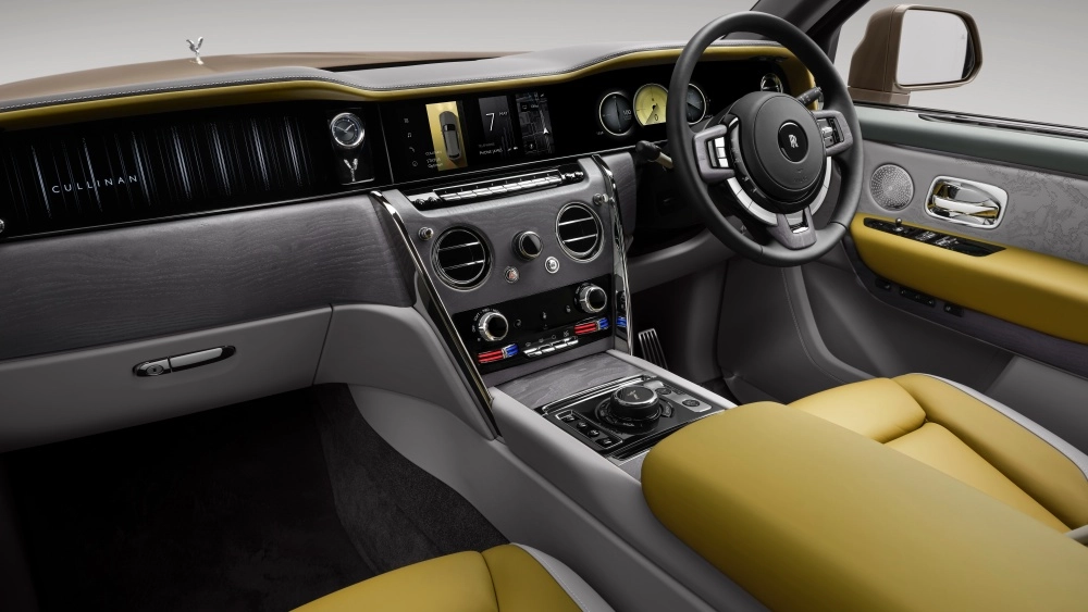 Rolls-Royce Cullinan Series 2 lüks SUV'un iç mekanında, gri ahşap kaplama, canlı sarı deri döşemeler ve parlak krom detaylarla süslenmiş, modern dijital ekranları, klasik bir analog saati ve kullanıcı dostu kontrol düğmelerini barındıran zarif ön konsol tasarımı net bir şekilde sergileniyor.