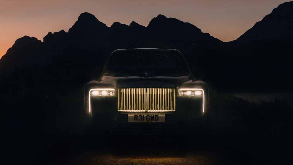 Gece, dağların karanlık siluetleri önünde duran, ön farları ve ızgarası parlak bir şekilde aydınlatılmış Rolls-Royce Cullinan Series 2 lüks SUV'un güçlü ve etkileyici ön tasarımı sergileniyor.
