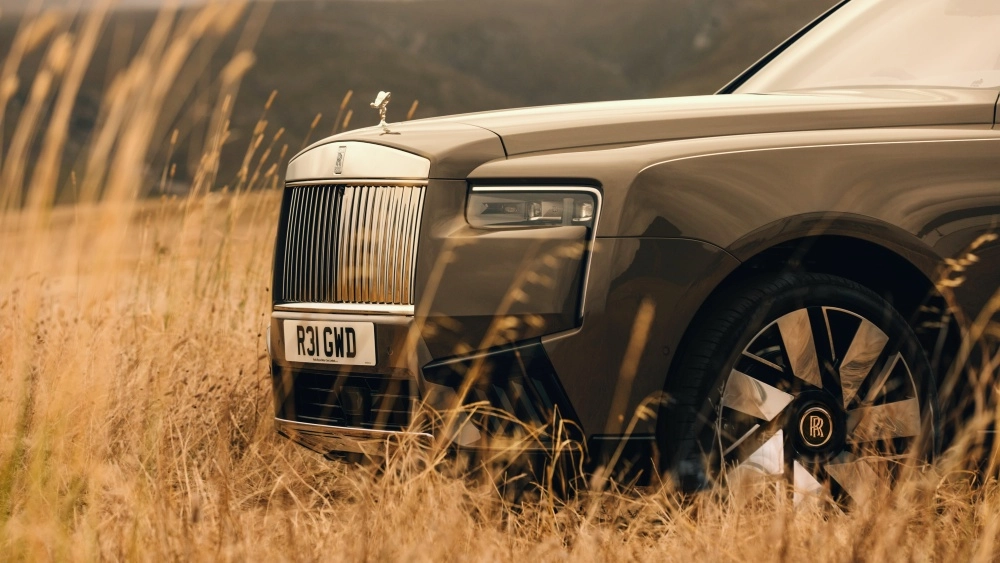 Yüksek, kuru otların arasından kısmen görünen Rolls-Royce Cullinan Series 2 lüks SUV'nin kendine özgü krom ızgarası, kaput üzerindeki Spirit of Ecstasy heykeli ve modern ön tasarım detayları, aracın şık duruşunu sergiliyor.