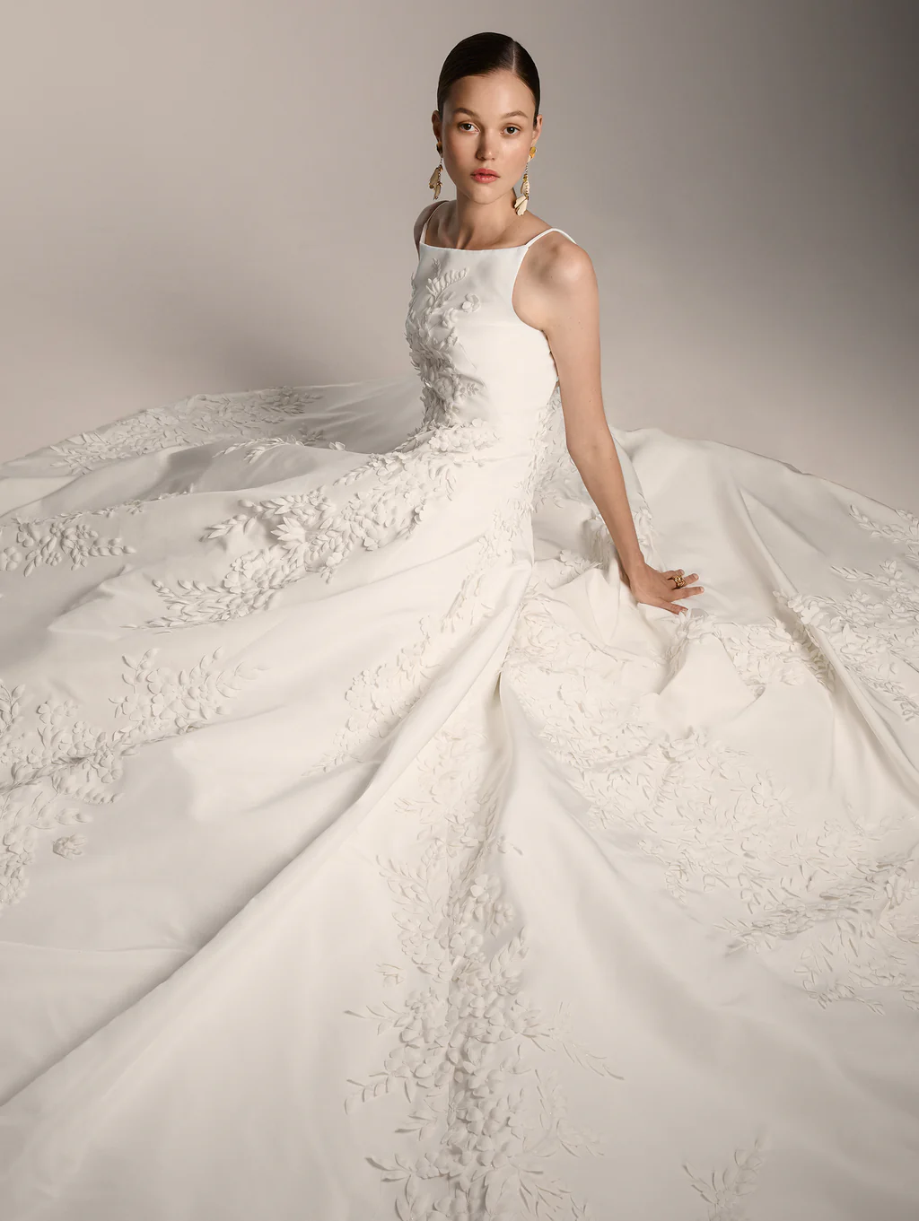 Beyaz, ince askılı ve kare yakalı bir Oscar de la Renta gelinlik giyen model, üç boyutlu çiçek ve yaprak aplikeleriyle bezeli kabarık etek kısmı etrafına yayılmış halde zarifçe oturuyor, romantik gelinlik estetiğini modern bir zarafetle harmanlıyor.
