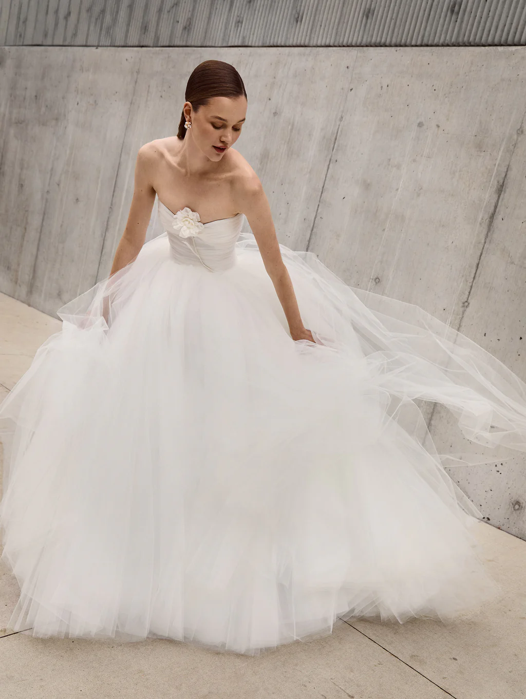 Oscar de la Renta imzalı, modern mimarinin beton duvarları önünde duran bu straplez romantik gelinlik modelinde, tülün katman katman oluşturduğu hacimli etek, korsajındaki tek ve zarif üç boyutlu çiçek detayıyla birleşerek masalsı bir zarafet sunuyor.