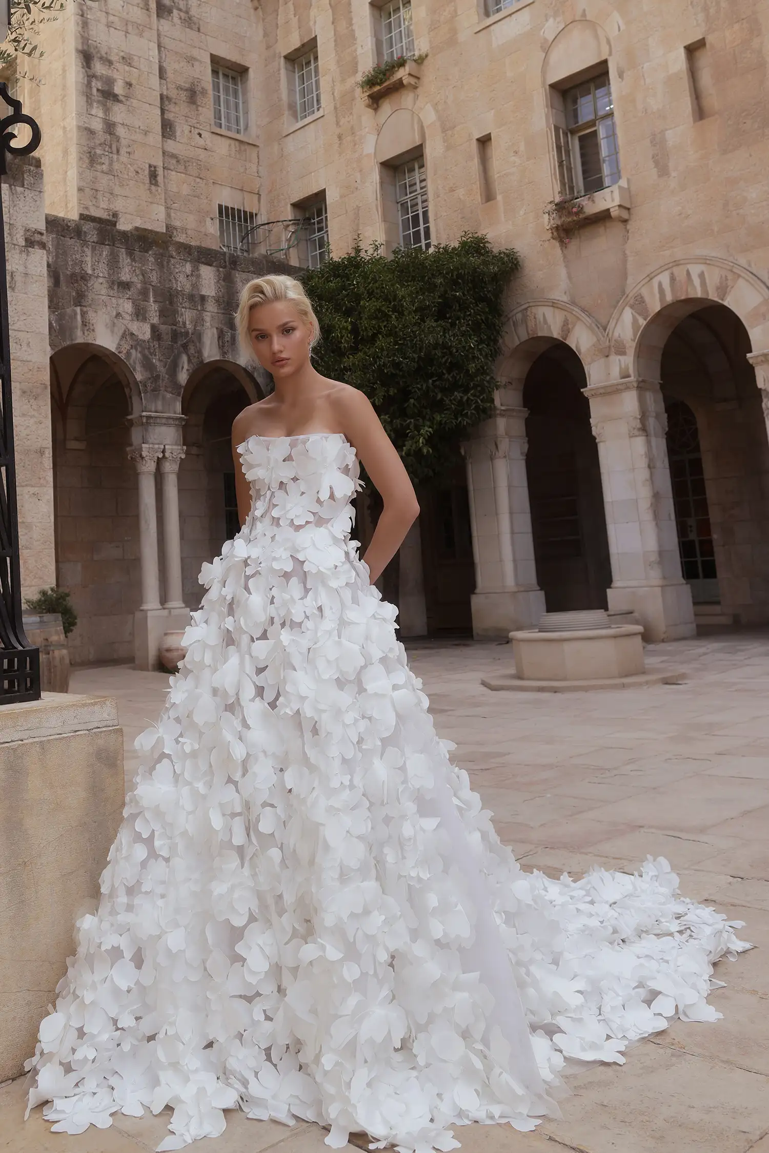 Kemerli taş binaların bulunduğu romantik bir avluda duran sarışın model, her yeri üç boyutlu çiçek işlemeleriyle bezeli, straplez korsajlı ve kabarık etekli görkemli bir romantik gelinlik modeli sunuyor.