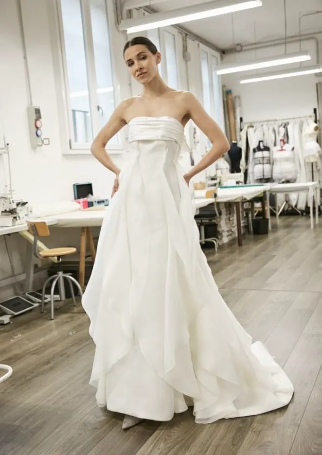 Bir moda atölyesi ortamında, elleri belinde poz veren zarif bir model, straplez, hacimli ve katmanlı drapelerle şekillendirilmiş etekleriyle dikkat çeken Peter Langner imzalı bir sade gelinlik modeli sunuyor; bu tasarım, 2026'nın modern gelinlik trendlerine uygun zamansız bir şıklık ve sanatsal bir duruşu bir araya getiriyor.