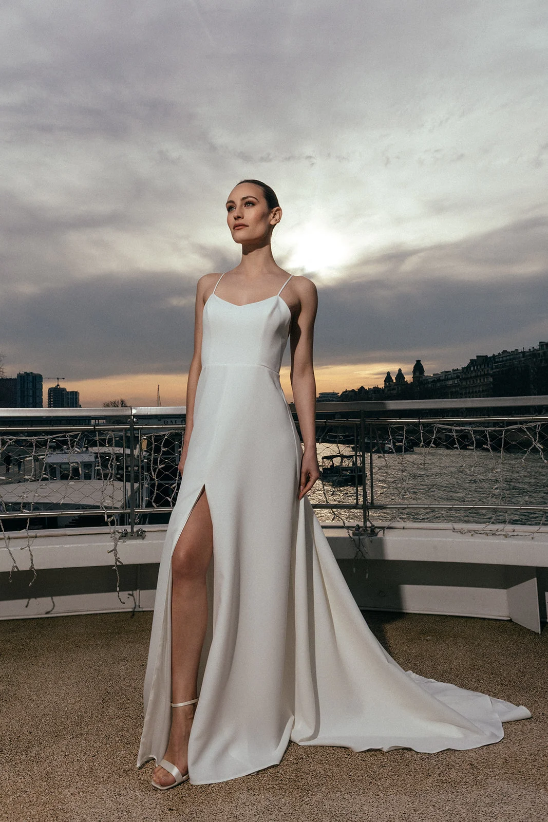 Cymbeline markasının sade gelinlik modellerinden, ince askılı, bacak yırtmaçlı ve uzun kuyruklu beyaz bir gelinlik giyen model, saçları topuz yapılmış halde, gün batımındaki bulutlu gökyüzü ve su manzarası önünde zarifçe duruyor.