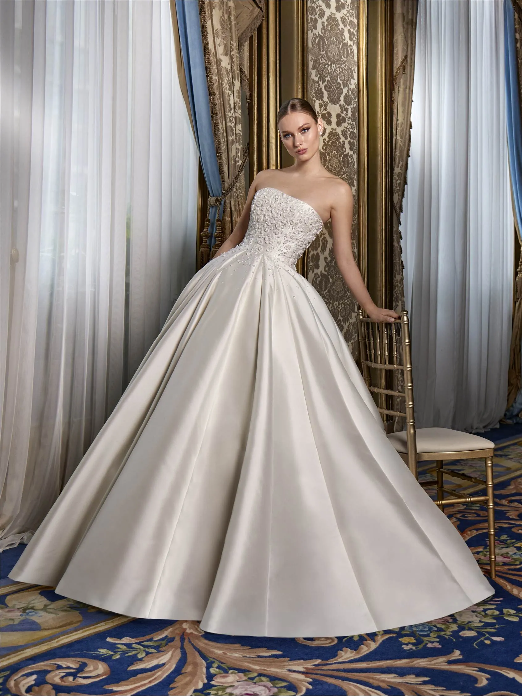 Pronovias markasına ait sade gelinlik modelleri arasındaki bu zarif tasarımda, göz alıcı saten kumaştan yapılmış kabarık eteği ve parlak taşlarla bezenmiş straplez korsesiyle model, işlemeli mavi halı serili lüks bir salonda altın rengi sandalyeye hafifçe yaslanarak poz veriyor.