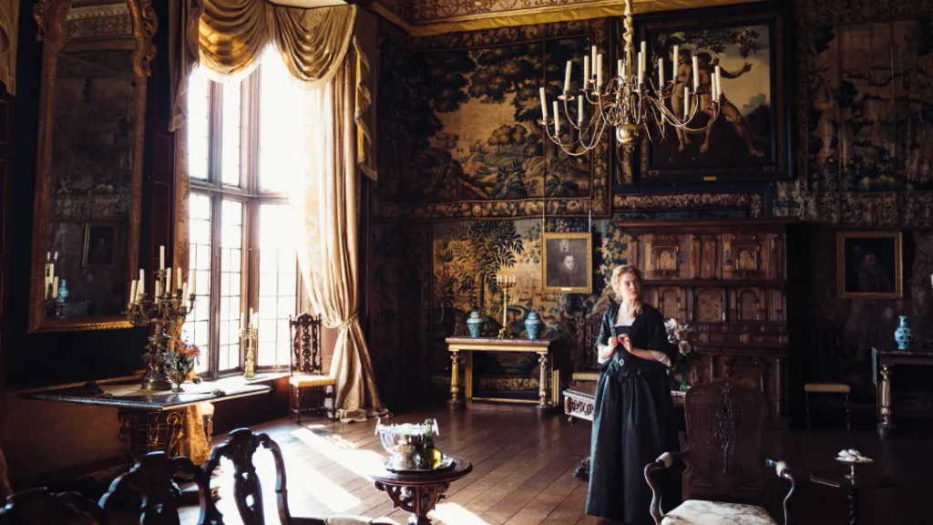 Sarayın Gözdesi (The Favourite) filminden bir sahne: Tarihi bir sarayın ihtişamlı salonunda, siyah elbise giymiş bir kadın karakter loş ışıkta ayakta duruyor. Duvarlar ağır işlemeli goblenlerle ve büyük portrelerle süslenmiş, yüksek pencerelerden gün ışığı süzülüyor. Ortam, filmin aristokratik ve dramatik atmosferini yansıtıyor.