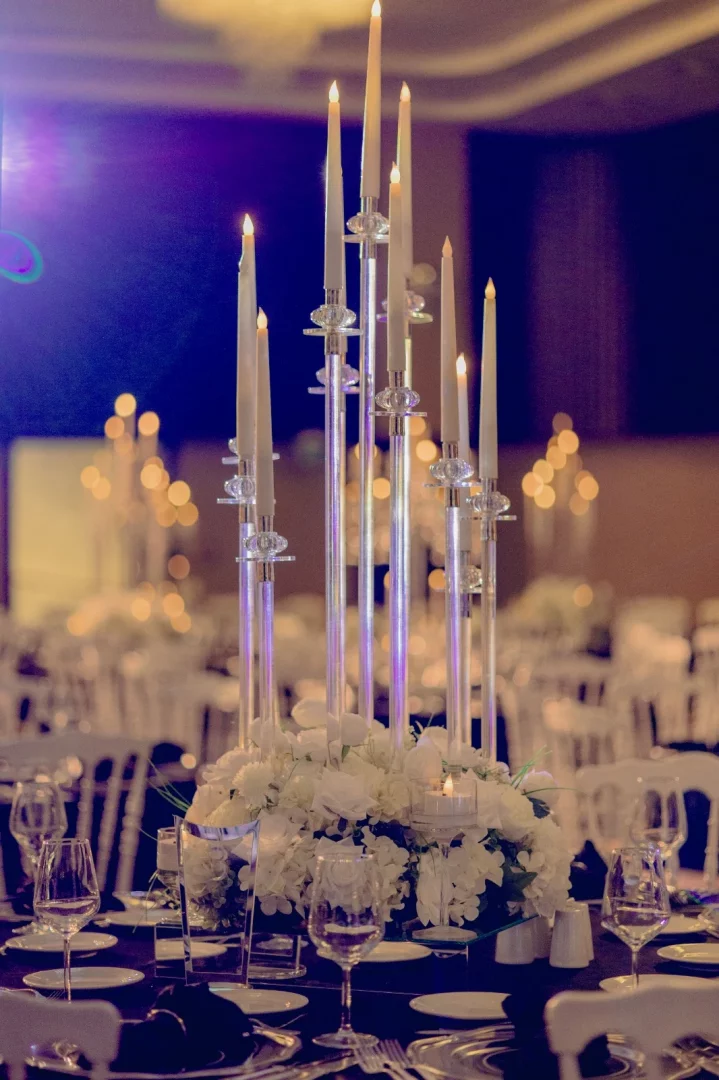 Sheraton Istanbul City Center'da düzenlenen gösterişli bir düğün kutlamasından, beyaz uzun mumlarla aydınlatılmış şık şamdanlar ve altındaki gür beyaz çiçek aranjmanıyla donatılmış, özenle kişiselleştirilmiş masa düzeninin yakın çekimi, arka planda bulanık misafir masaları ve sandalyelerle zarif bir ambiyans sergiliyor.