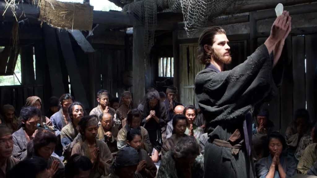 Silence (2016) filminden bir sahne: Rahip Rodrigues (Andrew Garfield), basit bir kulübede toplanmış Japon köylüler önünde ayin düzenliyor. Rahip, elinde kutsal ekmeği (hostiya) yukarı kaldırırken, etrafındaki köylüler dua eder şekilde başlarını eğmiş. Sahne loş, dramatik ışıkla aydınlatılmış ve inanç, umut ve sessizlik temalarını yansıtıyor.