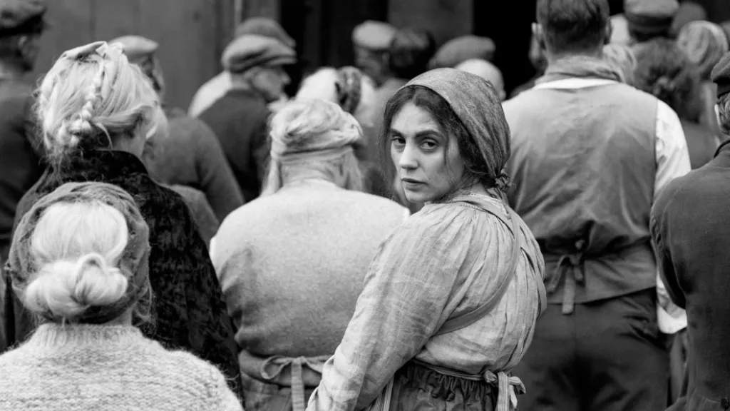 Şişli Kız filminden siyah-beyaz bir sahne: Kalabalık bir grup sırtı dönük şekilde beklerken, başörtülü bir kadın kalabalığın içinden geriye dönerek doğrudan kameraya bakıyor. Kadının yüz ifadesi güçlü ve sorgulayıcı. Sahne, dönemin toplumsal koşullarına dair etkileyici bir görsel atmosfer sunuyor.
