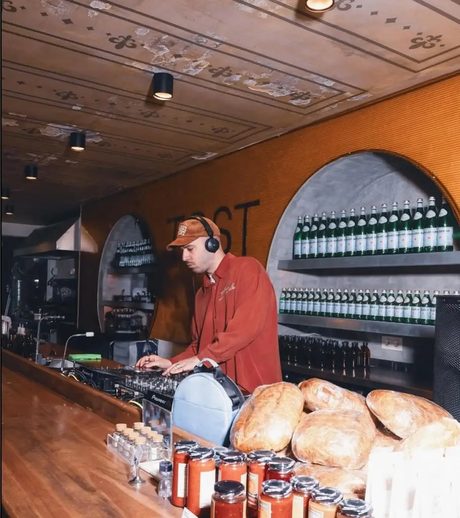 Alaçatı'da ahşap bir bar tezgahının arkasında kulaklık takmış bir DJ müzik çalarken, tezgahın üzerinde taze ekmekler, el yapımı sosları düşündüren kavanozlar ve baharatlıklar duruyor; duvarlarda görünen TOST yazısı ve arka plandaki şişe rafları, buranın tamamen organik tost çeşitleri sunan bir mekan olduğunu vurguluyor.