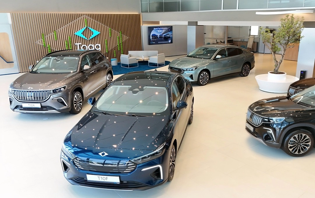 Aydınlık ve ferah bir Togg Deneyim Merkezi içindeki showroomda, önde parlak koyu mavi bir T10F sedan ve solunda mat gri bir T10X SUV öne çıkarken, arkada Togg logosu, rahat oturma alanları ve diğer yeni nesil modeller sergilenmektedir.