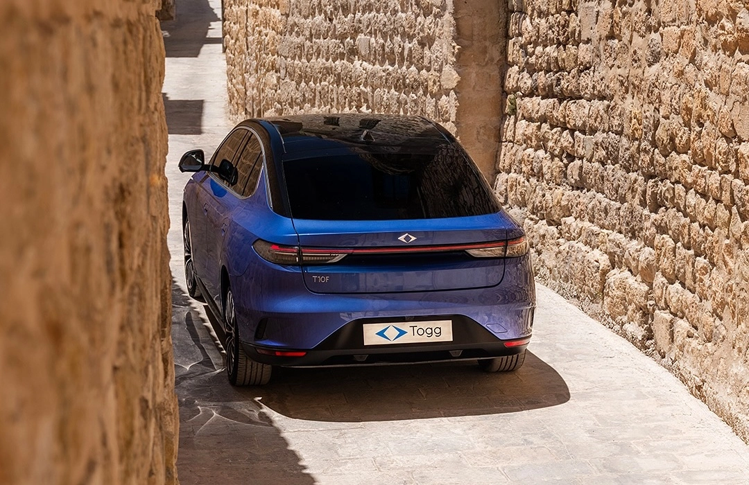 Mardin rengi Togg T10F elektrikli sedanın modern arka tasarımı, dar taş duvarlı tarihi bir sokağın içinde, gün ışığında aracın detaylarını ve çevresel uyumunu net bir şekilde gözler önüne seriyor.