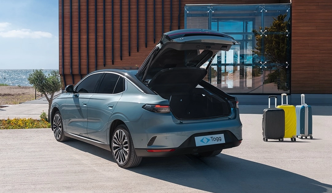 Deniz kenarında, modern bir binanın önünde, bagaj kapağı açık bir şekilde park edilmiş gri renkteki Togg T10F elektrikli sedan modeli, geniş bagaj hacmini vurgulayarak yanında duran üç farklı renkteki bavulla seyahat hazırlığını akılda canlandırıyor.