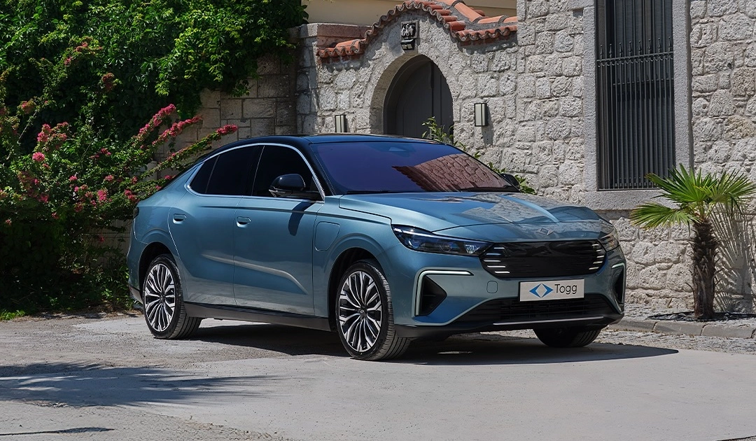 Taş duvarlı bir binanın ve kemerli girişinin önünde, yemyeşil bitkilerle çevrili, güneşli bir havada park etmiş parlak turkuaz renkteki Togg T10F elektrikli sedan modeli, Türkiye'nin gurur kaynağı yerli otomobil üretimini temsil ediyor.