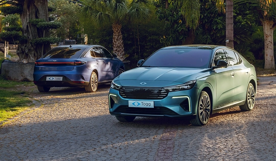 Çakıl taşlı bir zeminde, tropikal bitkiler ve ağaçlarla çevrili bir alanda duran, ön planda belirgin teal rengi ve şık tasarımıyla dikkat çeken Togg T10F elektrikli sedan modeli ile arka planda zarif lacivert tonundaki Togg T10F modelleri bu etkileyici araçları gözler önüne seriyor.